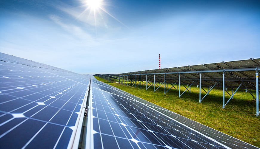 A view of a large solar farm with rows of solar panels under a bright blue sky. The sun shines intensely, casting reflections on the panels. Green grass is visible beneath the panels, and in the background, a tower with red and white stripes can be seen.