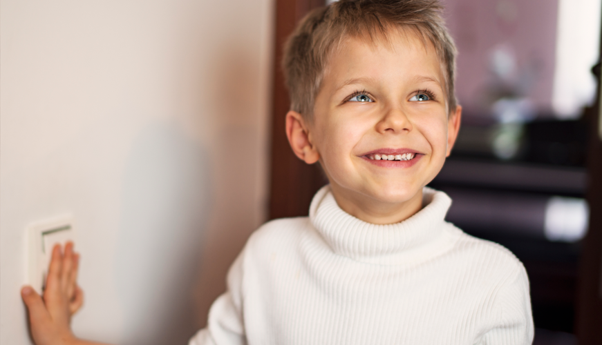 A young child with short hair, dressed in a white turtleneck, stands indoors smiling and looking to the side. They are touching a light switch on a wall. The background is softly blurred.