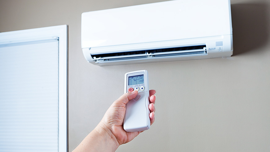 A hand holding a remote control is pointing at a wall-mounted air conditioning unit. The background shows a partial view of a window with closed blinds.