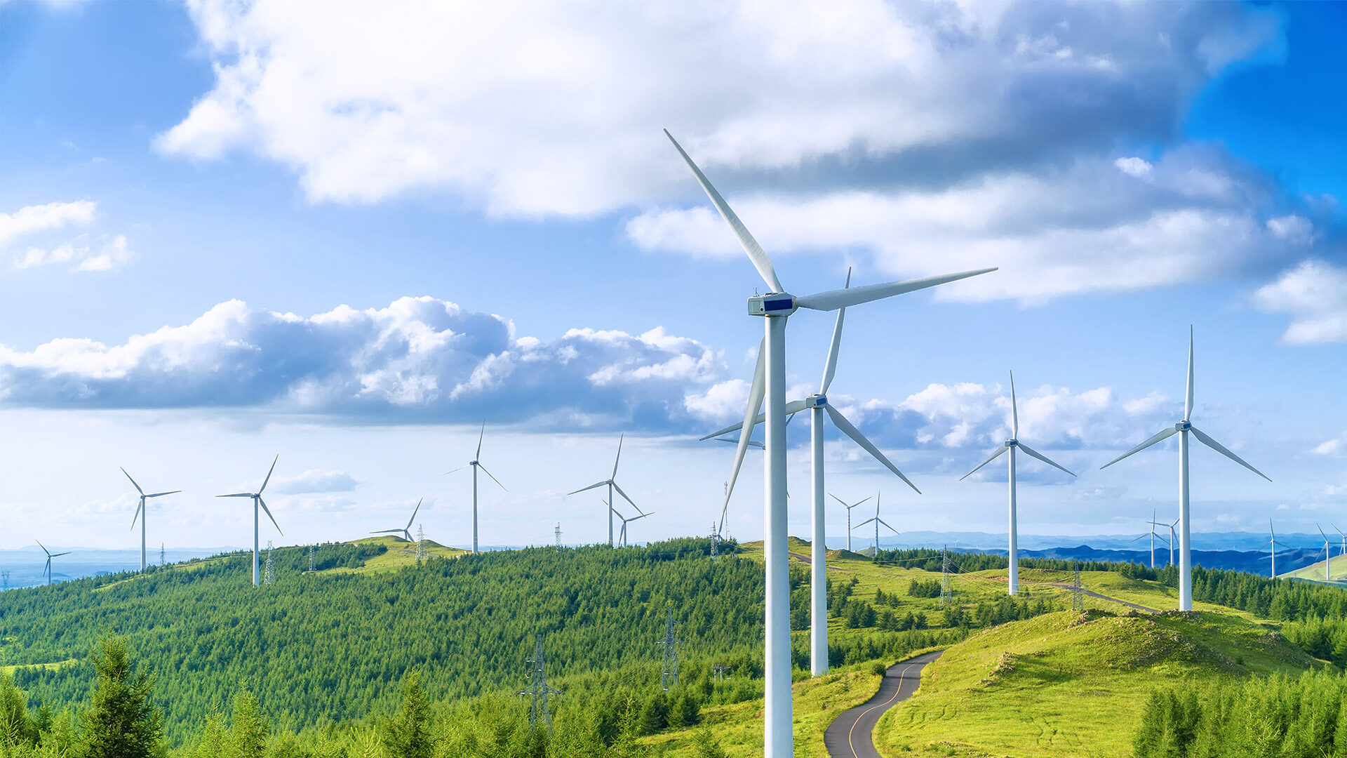 A panoramic view of several wind turbines on a green hill under a partly cloudy blue sky. The wind turbines are surrounded by lush vegetation, and a winding road is visible weaving through the hills.