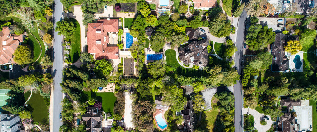 Aerial view of a suburban neighborhood featuring tree-lined streets and large houses with gardens and swimming pools. The area is lush with greenery, creating a vibrant and organized landscape.